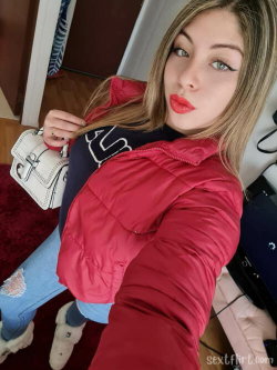 A woman with blonde highlights takes a selfie in a red puffer jacket, ripped jeans, and white boots, holding a small