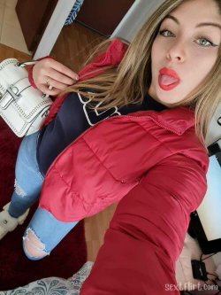 A woman with long brown hair and red lipstick sticks her tongue out in a playful selfie, wearing a red puffer jacket,