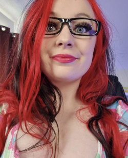 Woman with vibrant red hair and black-framed glasses, wearing a colorful patterned top, smiling in a close-up selfie.