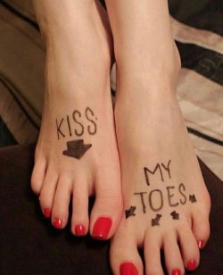 Close-up of bare feet with red toenails and handwritten text 'KISS MY TOES' on the soles, resting on a dark surface.