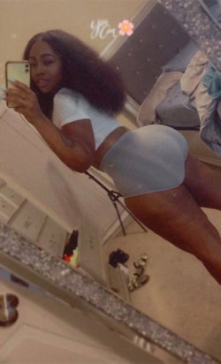 A woman with long curly hair takes a mirror selfie in a white crop top and light gray shorts, showcasing a curvy figure