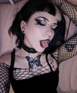 A woman with dark hair and bold makeup lies on a bed, wearing black fishnet sleeves and a choker, with a butterfly