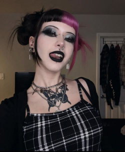 Person with black and pink hair, dark makeup, multiple piercings, and a chest tattoo wearing a plaid top indoors.