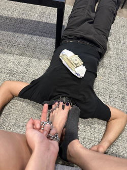 Person lying on carpet in black pants and shirt, money tucked into waistband, feet chained and held by another hand.