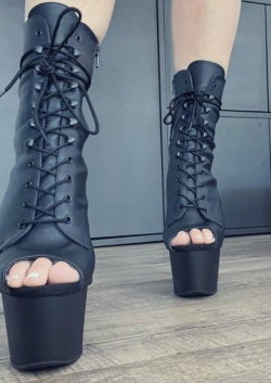 Close-up of legs wearing black lace-up platform boots with open toes, standing on a wooden floor.