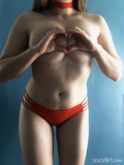 Curvy woman in red thong and choker covering her breasts with hands forming a heart shape against a blue background.