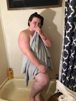 A curvy person with dark hair stands in a bathtub, wrapped in a towel, holding it near their face with one hand on