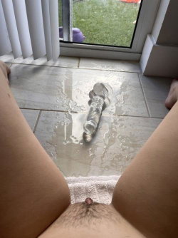 Close-up of a person's lower abdomen and thighs, with a glass sex toy and liquid on tiled floor near a window.