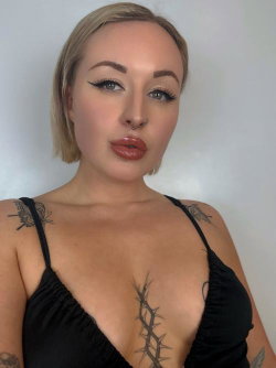 Blonde woman with short hair, wearing a black top, visible tattoos on chest and shoulders, posing against a plain
