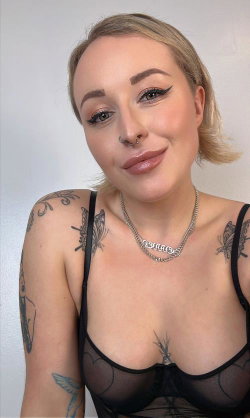 Blonde woman with short hair, wearing a black lace bra, visible tattoos on shoulders and chest, smiling at the camera.