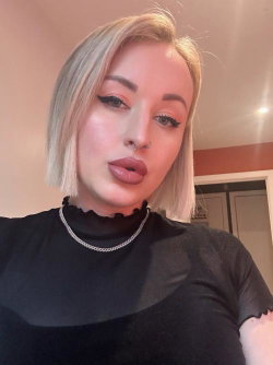 Blonde woman with short bob, wearing a black ruffled top and silver chain necklace, taking a selfie indoors.