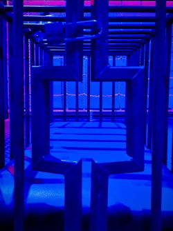 A dimly lit maze with blue and pink neon lighting, featuring metal frames and reflective surfaces.