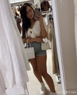 Brunette woman in white crop top and denim shorts takes mirror selfie in clothing store, holding phone and shopping bag.