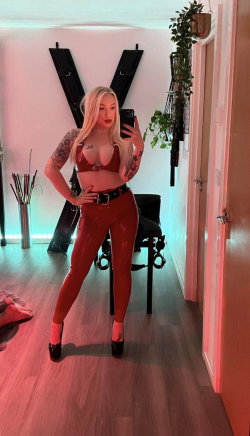 Blonde woman in red latex outfit takes mirror selfie, showing tattoos on arms and torso, standing in room with plants