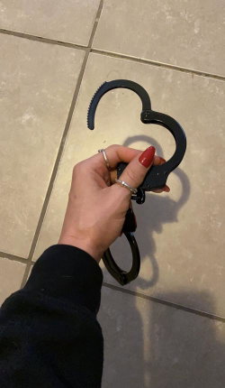 A hand with red nails and rings holds black handcuffs over a tiled floor, wearing a dark long-sleeve top.