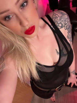 Blonde woman with red lipstick and nose ring, wearing a black mesh top, showing tattoos on her shoulder and chest in a