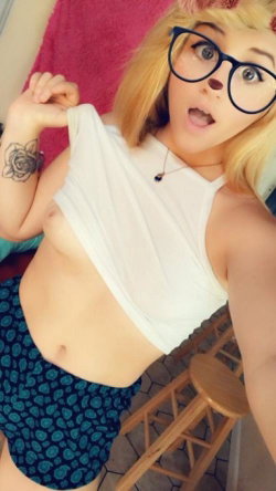 Blonde woman with glasses and a rose tattoo takes a selfie, lifting her white crop top to expose her midriff while
