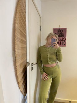 Blonde woman in olive green crop top and matching pants takes a mirror selfie in a room with a door and wall art.