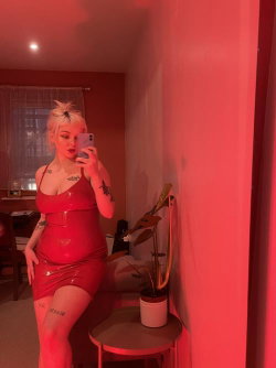 A woman with short blonde hair and visible tattoos takes a mirror selfie in a red latex dress, standing in a warmly lit