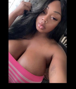 A Black woman with long dark hair and a nose ring poses topless in a pink striped garment, holding her hair while