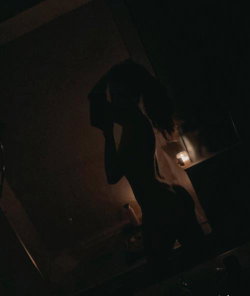 Silhouette of a person with long hair standing in dim lighting, possibly near a candle-lit surface, wearing dark