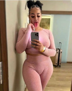 Curvy woman in pink mesh two-piece outfit takes a selfie, sticking tongue out while leaning against a wall indoors.