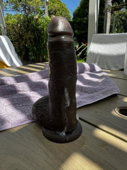 A dark brown, textured sex toy with a flared base sits on a wooden table outdoors, next to a purple towel with greenery