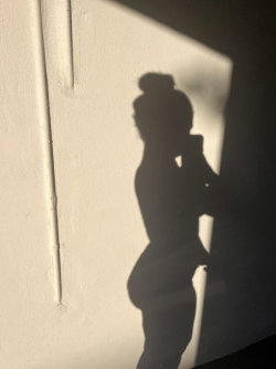 Silhouette of a woman with hair in a bun, holding a phone, standing against a white wall in shadow.