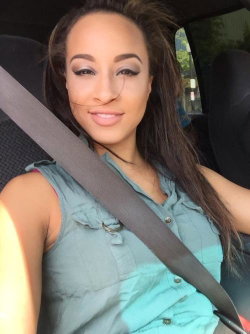 A woman with long dark hair and makeup smiles in a car selfie, wearing a sleeveless teal top with a seatbelt across her
