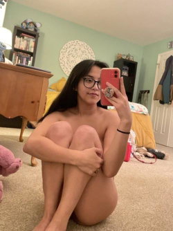 A woman with long dark hair and glasses sits nude on the floor, taking a mirror selfie in a bedroom with green walls