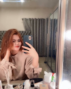 A woman with long reddish-brown hair takes a mirror selfie in a beige blouse, sitting at a vanity with beauty products