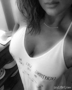 Close-up of a woman in a white tank top with text, wearing a delicate necklace, indoors with soft lighting.