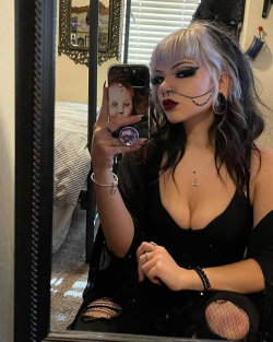 A woman with silver and black hair, dramatic makeup, and a black outfit takes a mirror selfie in a bedroom setting.