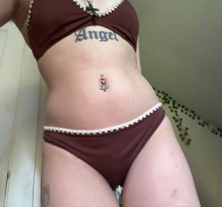 Curvy woman in brown lace-trimmed lingerie with visible tattoos on abdomen and thigh, posing indoors.