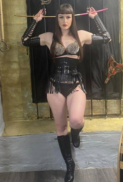 A woman with long dark hair and bangs wears a studded bra, black leather corset, and thigh-high boots, holding sticks
