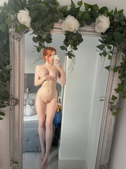Redheaded woman takes mirror selfie nude, covering breasts with hands, standing in bedroom with floral-decorated mirror.