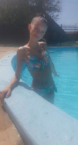 A slim woman in a colorful floral bikini poses at the edge of a swimming pool, smiling softly with her hair tied up.