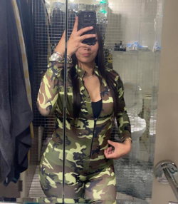 Woman in camouflage jumpsuit takes mirror selfie in bathroom, long dark hair, visible black top underneath.