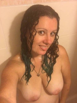 A woman with curly dark hair and green-tipped ends smiles in a shower, topless and wearing a necklace.