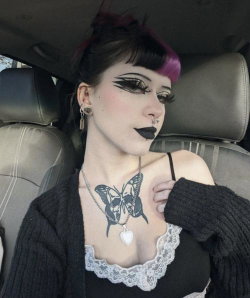 A woman with purple-tipped hair and dramatic makeup sits in a car, wearing a black lace top and cardigan, showing a