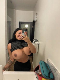 A smiling woman with long dark hair takes a mirror selfie in a bathroom, wearing a black mesh top and black bottoms,