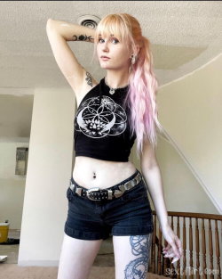 A slim woman with pink-tipped blonde hair wears a black crop top and denim shorts, posing indoors with visible tattoos