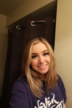 Blonde woman with shoulder-length hair smiles in a selfie, wearing a purple graphic tee in front of a dark curtain.