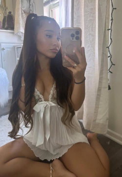 A woman with long dark hair in a high ponytail takes a mirror selfie, wearing a white lace-trimmed top and sitting