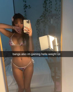Asian woman with bangs in floral bikini takes mirror selfie in bedroom, hand near mouth, holding phone.