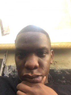A Black man with a shaved head and dark skin poses in a dimly lit room, hand resting on his chin, wearing a dark shirt.