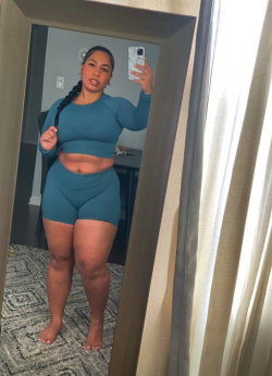 A curvy woman with dark hair in a braid takes a mirror selfie in a teal long-sleeve crop top and matching shorts,