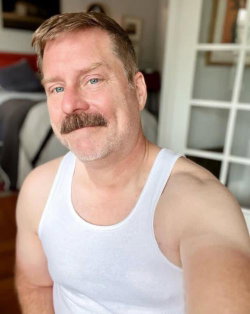 A man with a mustache and short hair wears a white tank top, taking a selfie indoors with soft lighting.