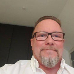 A man with glasses and a gray beard wearing a white shirt, taking a selfie indoors.