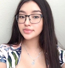 A young woman with long dark hair and glasses wears a floral top, posing closely against a plain wall in a selfie-style